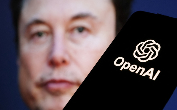ภาพเบลอใบหน้า Elon Musk อยู่ด้านหลัง ขณะโลโก้ OpenAI ปรากฏชัดบนหน้าจอโทรศัพท์ด้านหน้า สื่อถึงความสัมพันธ์ที่ตึงเครียดระหว่างผู้เล่นรายใหญ่ในวงการ AI